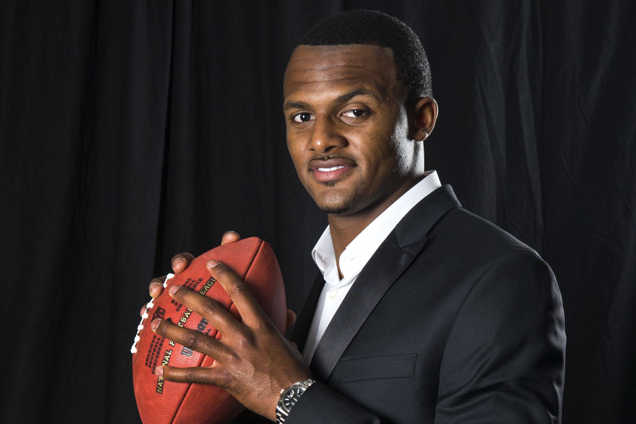Deshaun Watson 桌布 壁紙 — 一位自信的非裔美國男子穿著黑色西裝，在深色背景前拿著一個紅色橄欖球。