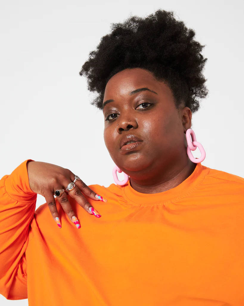 Black Girl 桌布 壁紙 — 一位自信的非裔髮型女性，在純白背景前穿著大膽的橙色衣服和超大粉紅色耳環。