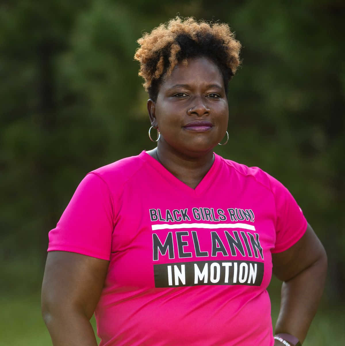 黑人女性 桌布 壁紙 — 一位自信的黑人女性，捲髮，穿著亮粉紅色「Black Girls Run Melanin in Motion」T恤，背景是模糊的綠色森林。