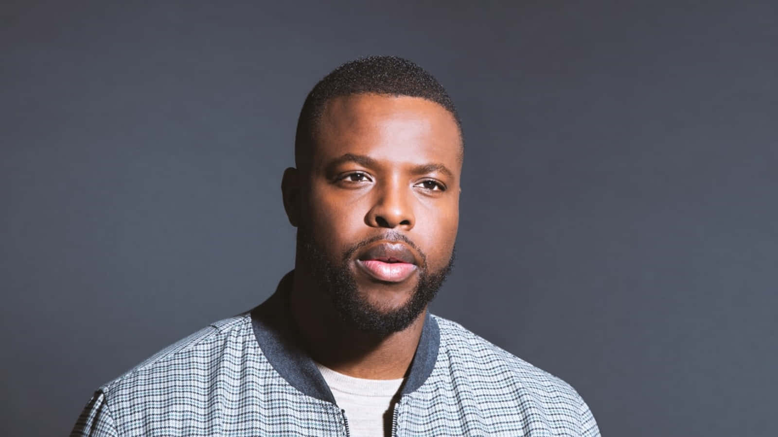 Winston Duke 桌布 壁紙 — 一位自信的黑人鬍鬚男穿著格紋外套，背景為深灰色攝影棚。
