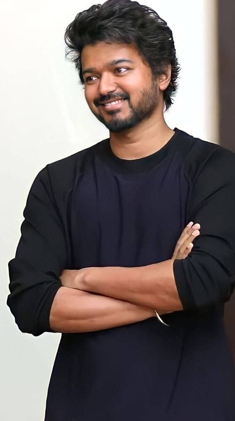 vijay HD 桌布 壁紙 — 一位自信的黑髮鬍鬚男士，穿著黑色襯衫，交叉雙臂，在純色背景前微笑。
