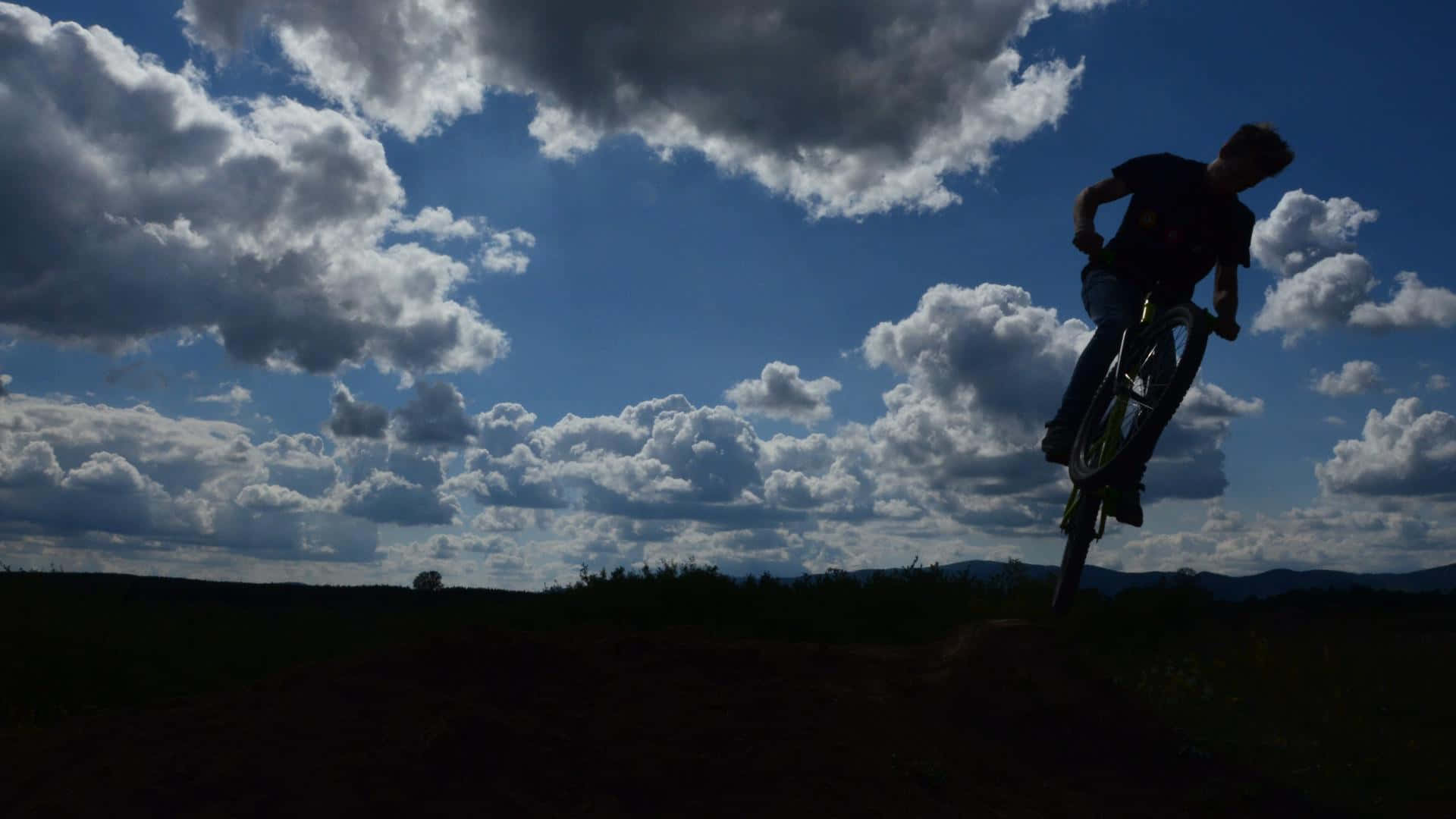 Mtb 桌布 壁紙 — 一位自行車手的剪影在戲劇性的多雲天空下，於開闊田野中跳過一個土丘。