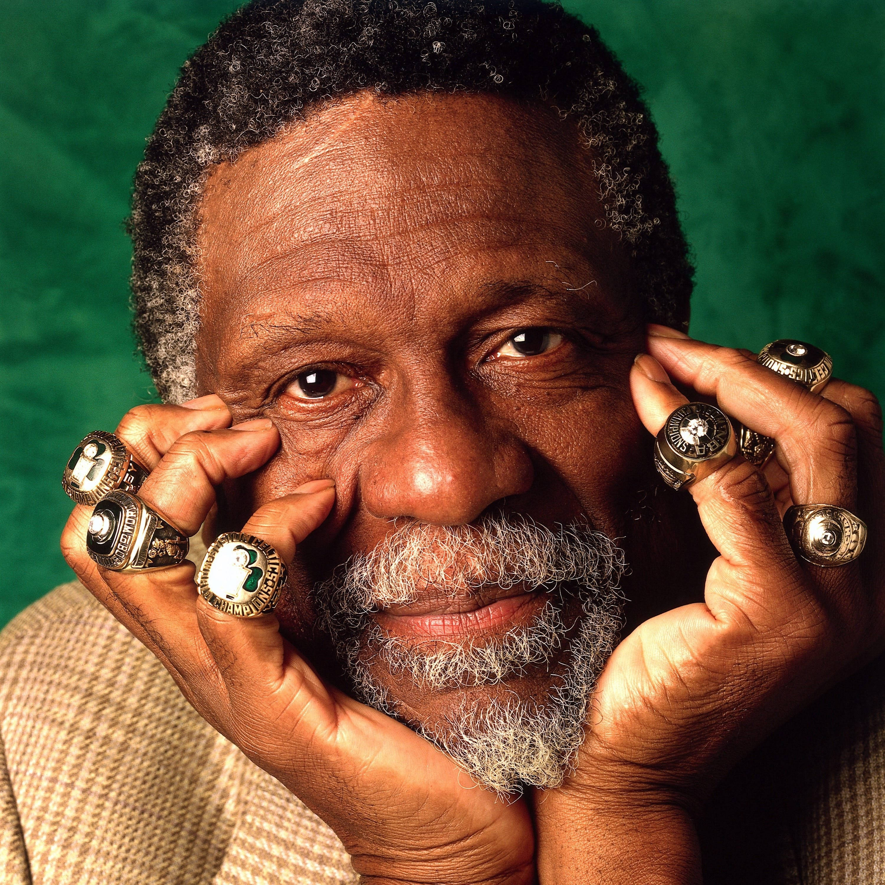 Boston Celtics 桌布 壁紙 — 一位自豪的退休運動員，留著灰色鬍鬚，戴著多枚冠軍戒指，在綠色背景前溫暖地微笑。