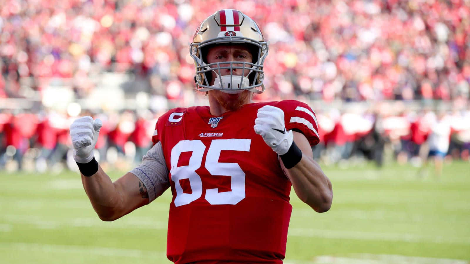 George Kittle 桌布 壁紙 — 一位舊金山四九人隊的足球員，身穿紅色85號球衣，在陽光明媚的球場上高舉拳頭慶祝。
