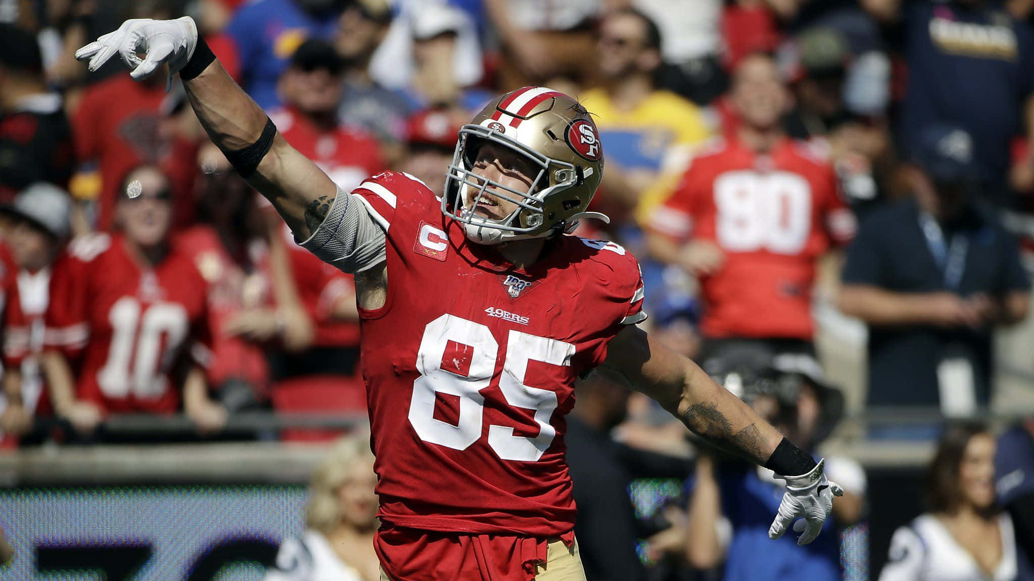George Kittle 桌布 壁紙 — 一位舊金山四九人隊的足球員，身穿紅色球衣85號，在陽光明媚的比賽日張開雙臂慶祝。