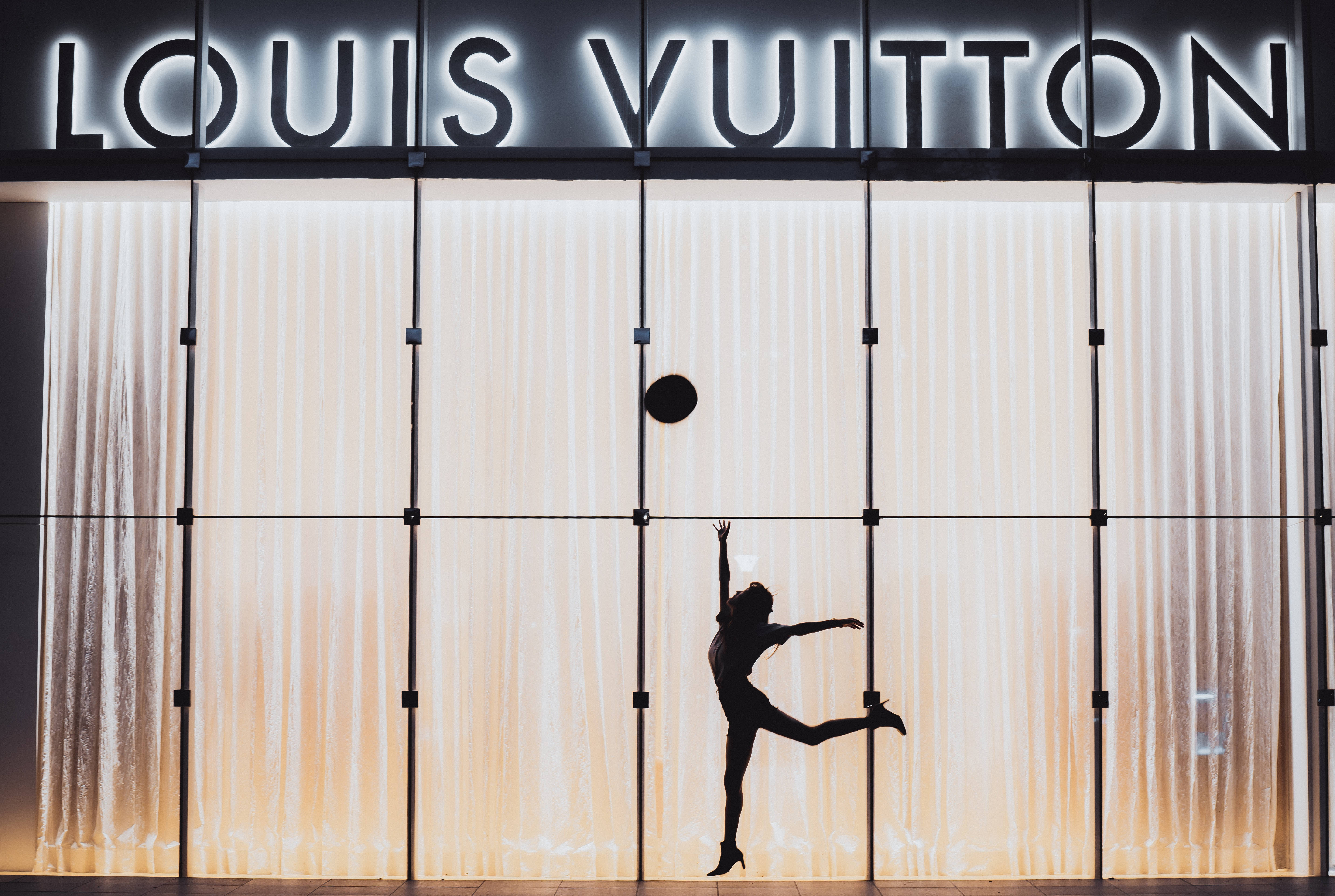 路易威登 桌布 壁紙 — 一位舞者的剪影在發光的 Louis Vuitton 店面（帶有垂直燈板）前優雅地跳躍。