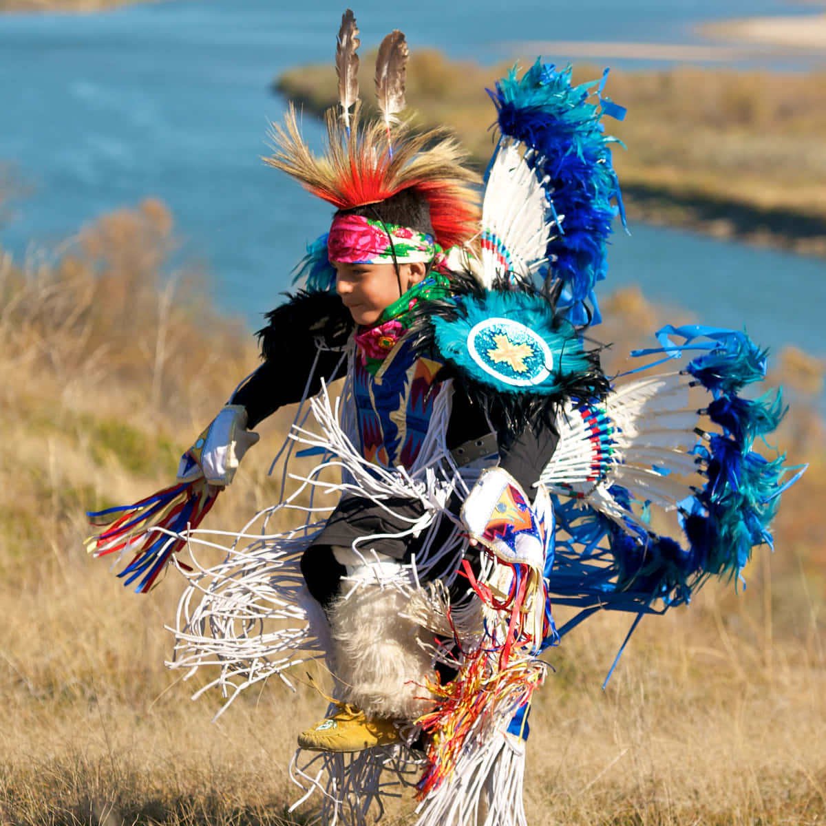 Native American 桌布 壁紙 — 一位舞者身穿鮮豔的傳統服飾，飾有藍色羽毛和彩色流蘇，在河邊充滿活力地表演。