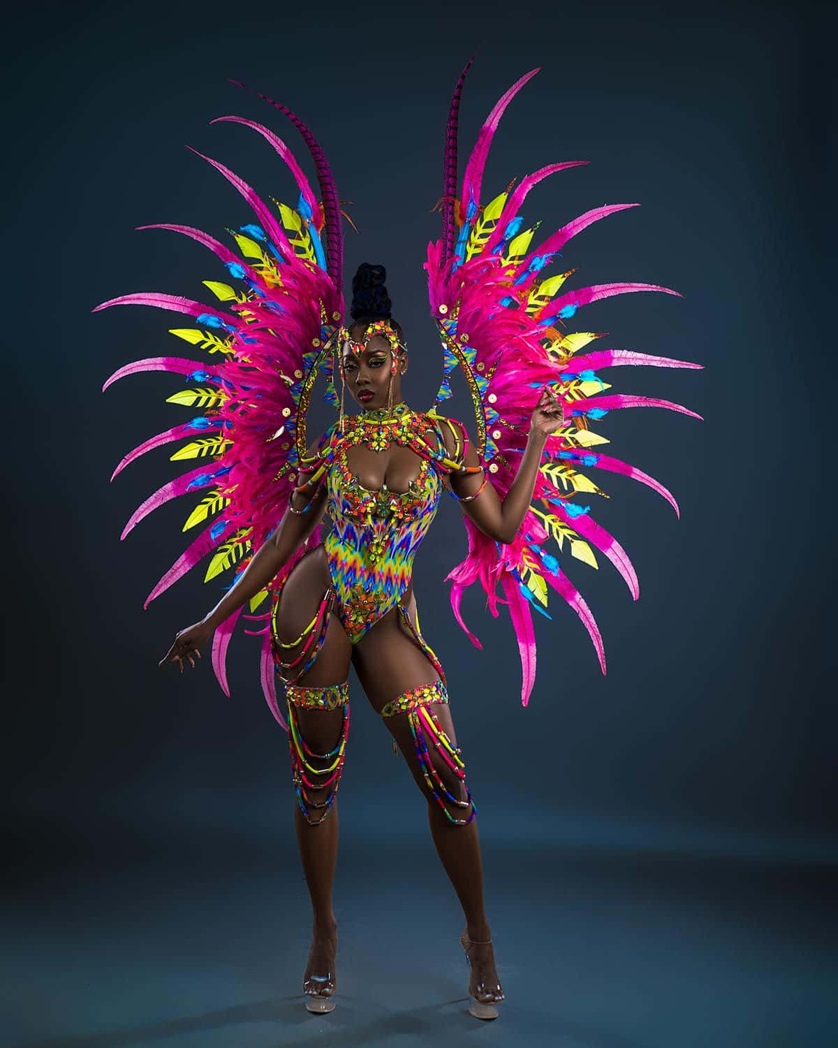 Carnival 桌布 壁紙 — 一位舞者身穿鮮豔的彩虹羽毛服裝，搭配巨大的粉紅色翅膀，在黑暗的攝影棚背景下自信地擺姿勢。