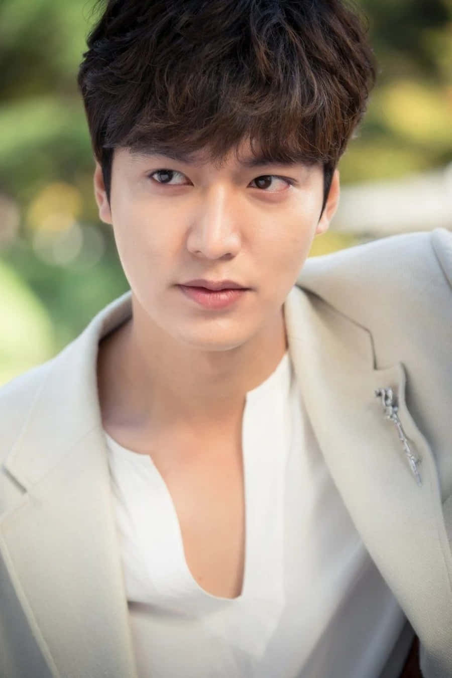 Lee Min Ho 桌布 壁紙 — 一位英俊男士身穿淺米色西裝外套和白色襯衫，在柔和自然光線和模糊綠色樹葉背景下專注地凝視。