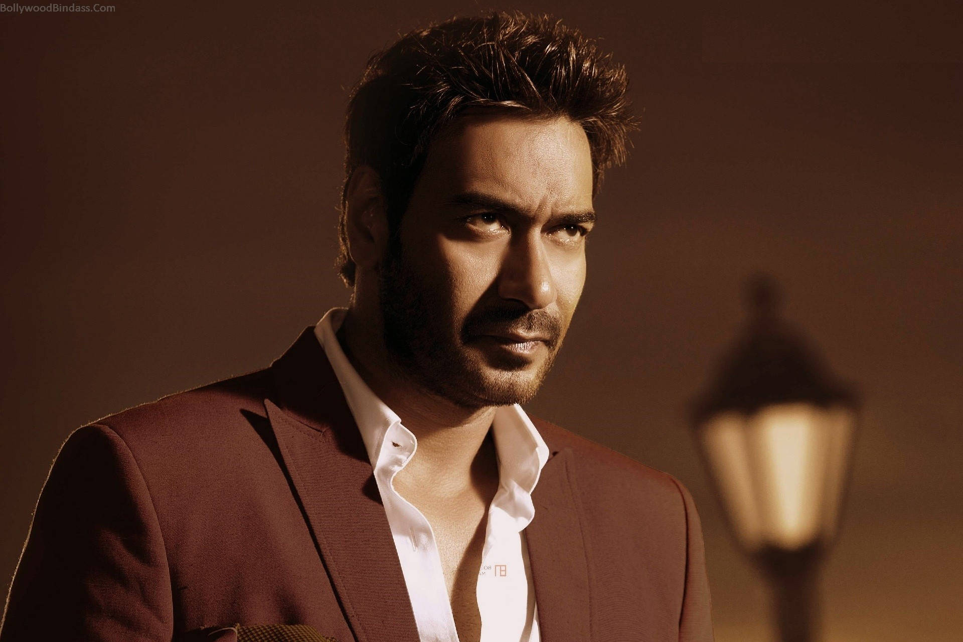 ajay devgn 桌布 壁紙 — 一位英俊男士身穿酒紅色西裝外套和白色襯衫，在溫暖的金色燈光下，背景中有一盞模糊的復古路燈，他凝視著鏡頭。