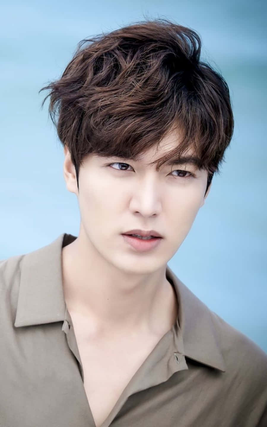Lee Min Ho 桌布 壁紙 — 一位英俊的年輕男子，一頭凌亂的棕色頭髮，眼神專注，身穿米色襯衫，背景是柔和的藍色。