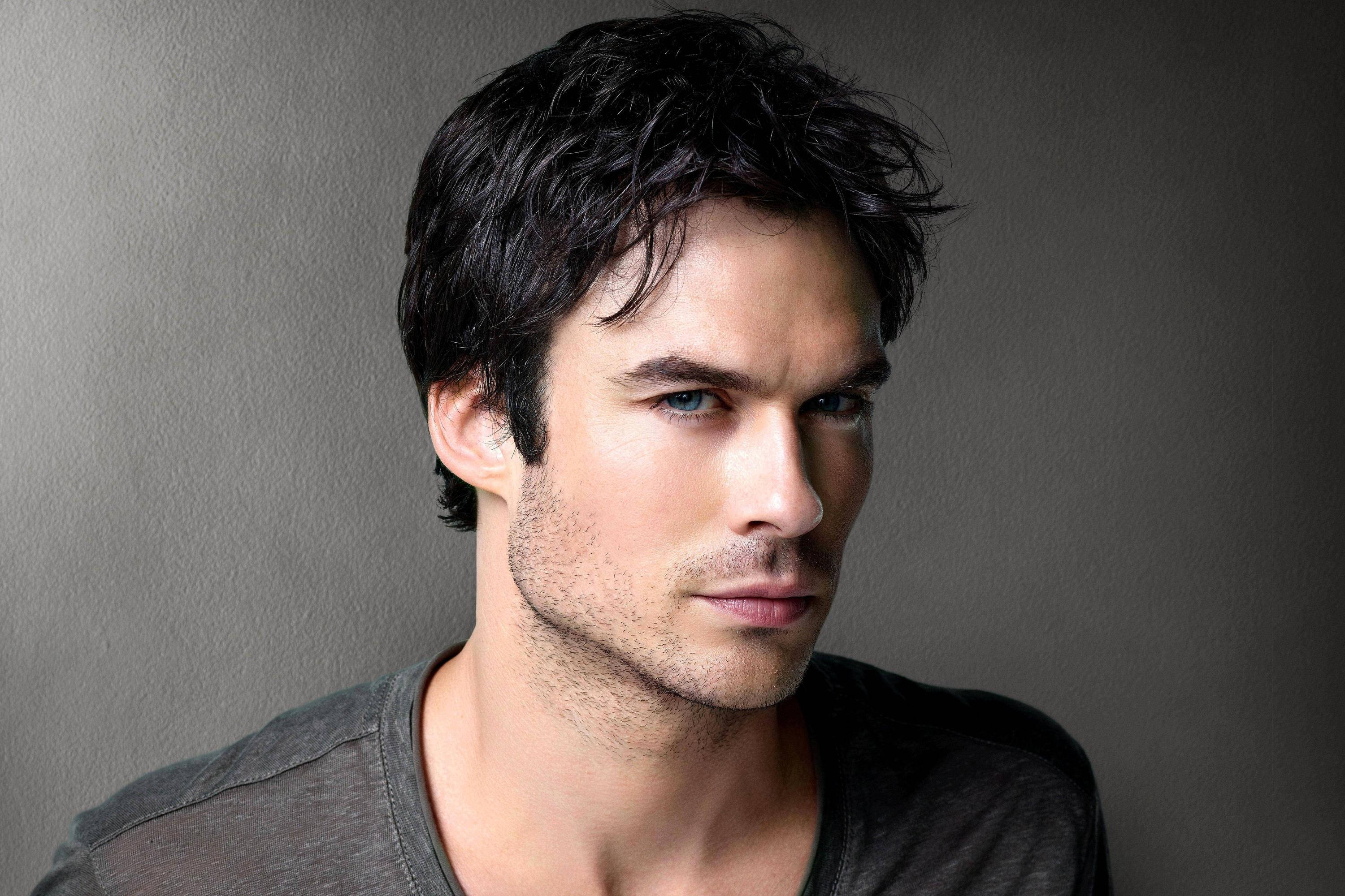 Damon Salvatore 桌布 壁紙 — 一位英俊的男人，深色凌亂的頭髮和銳利的藍眼睛，在柔和的灰色工作室背景下專注地凝視。