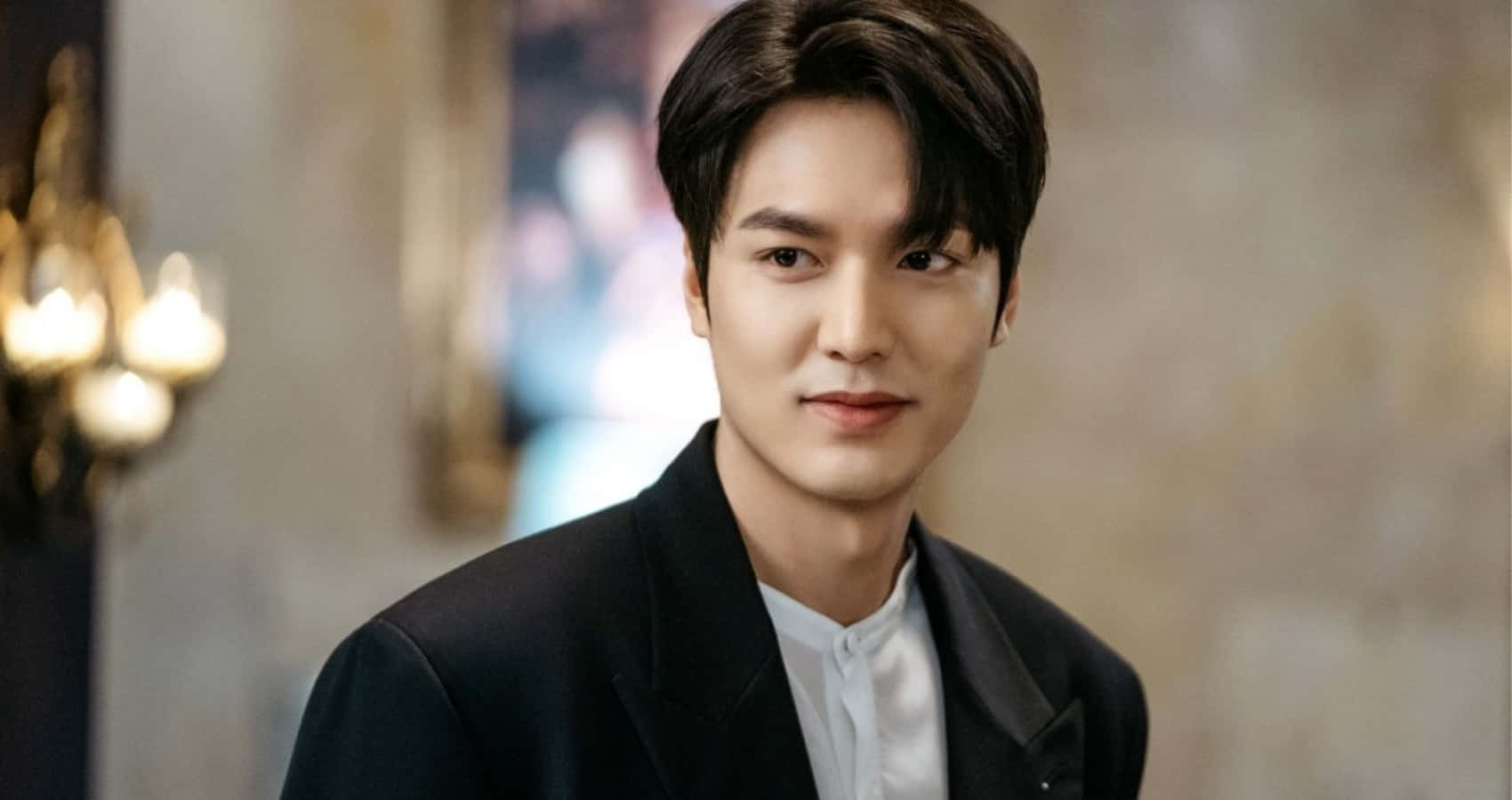 Lee Min Ho 桌布 壁紙 — 一位英俊的男士穿著黑色西裝外套和白色襯衫，在柔和光線的優雅室內環境中平靜地凝視著鏡頭。