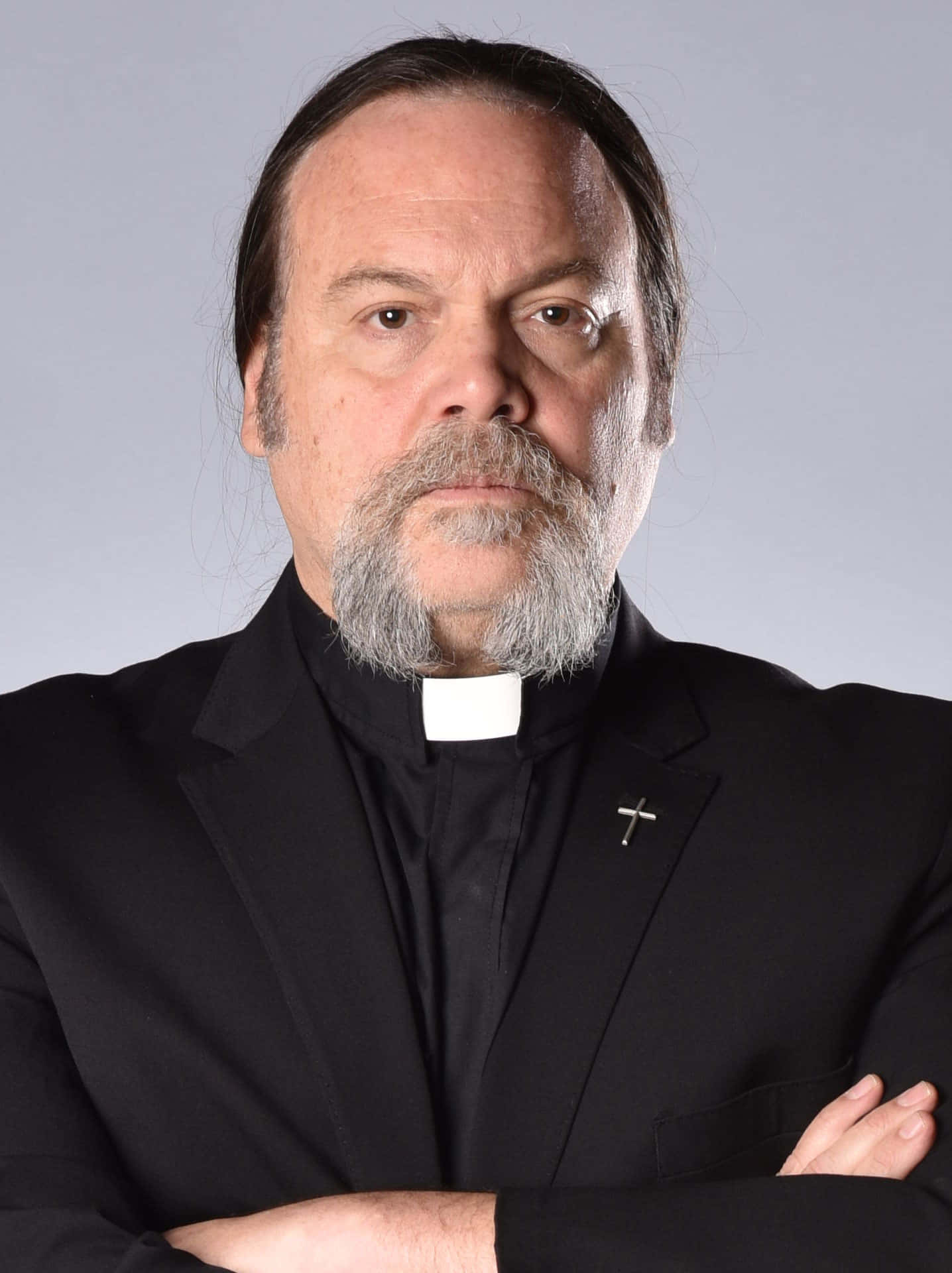 Vincent D'onofrio 桌布 壁紙 — 一位莊嚴的中年神父，留著灰色鬍鬚，雙臂交叉，穿著黑色神職人員領和一個小十字架別針，背景是純灰色。