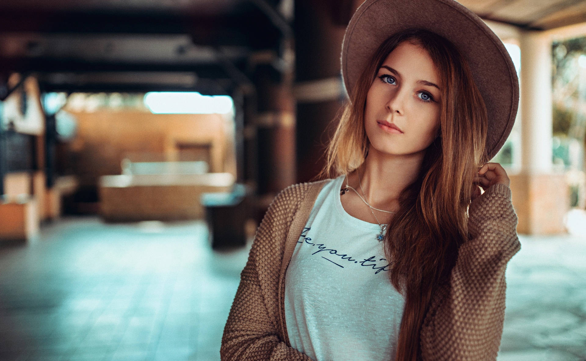 casual 桌布 壁紙 — 一位藍眼睛、長棕髮的年輕女性戴著寬邊帽和質感開襟衫，在柔和光線的室內空間若有所思地凝視鏡頭。