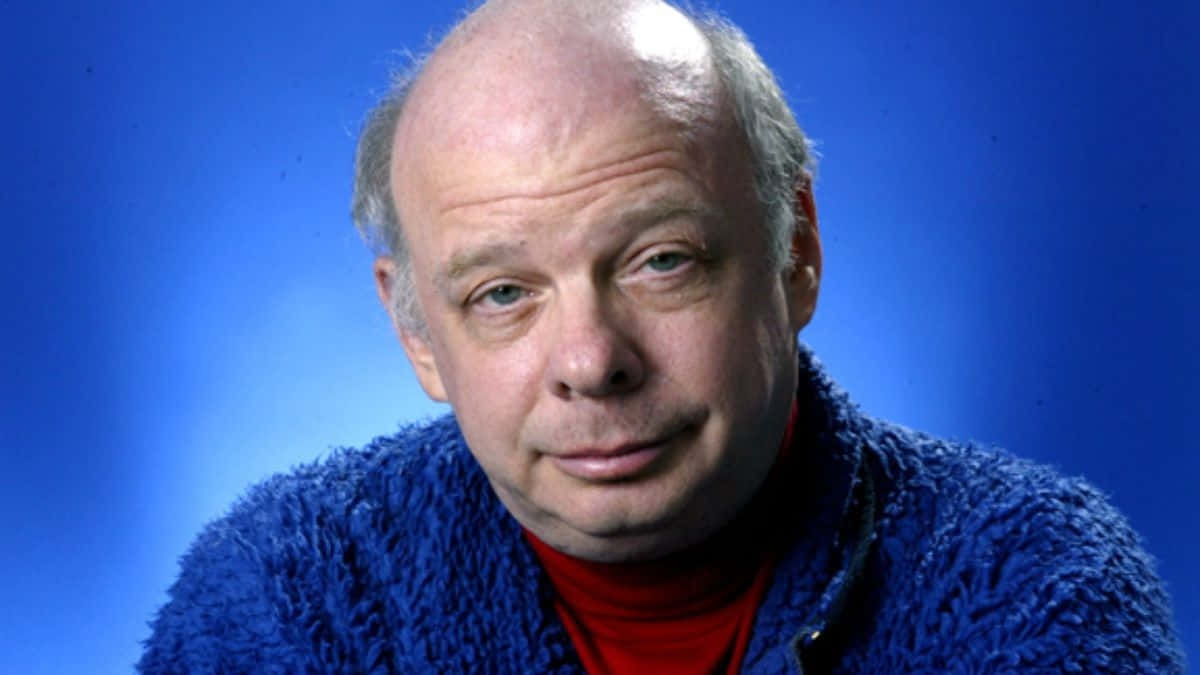 Wallace Shawn 桌布 壁紙 — 一位藍眼睛的光頭男士，在純藍色攝影棚背景前，穿著紅色高領毛衣外搭一件毛茸茸的藍色毛衣。