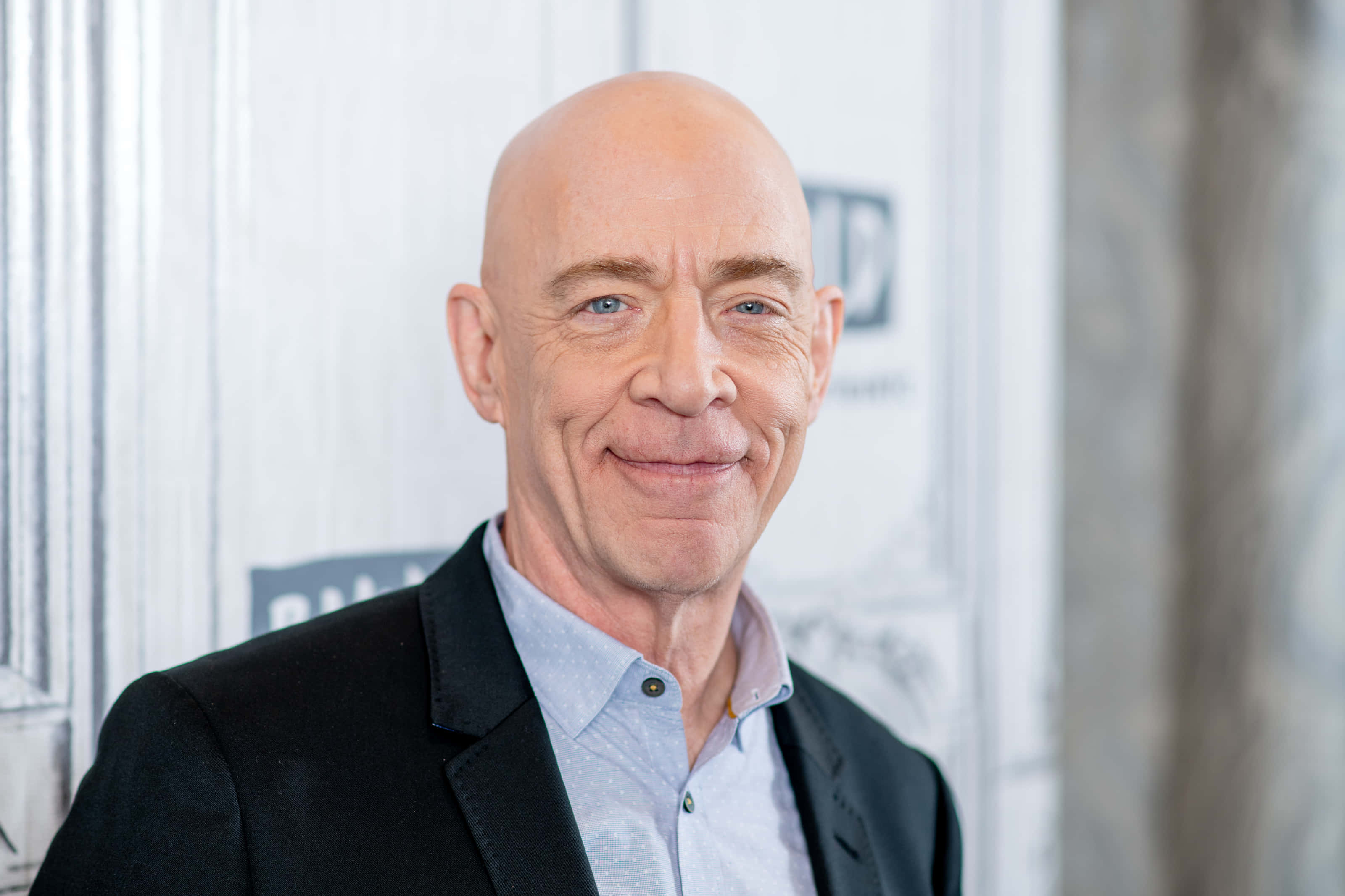 J.k. Simmons 桌布 壁紙 — 一位藍眼睛的光頭男士，身穿黑色西裝外套和淺藍色襯衫，在模糊的白色背景下溫暖地微笑。