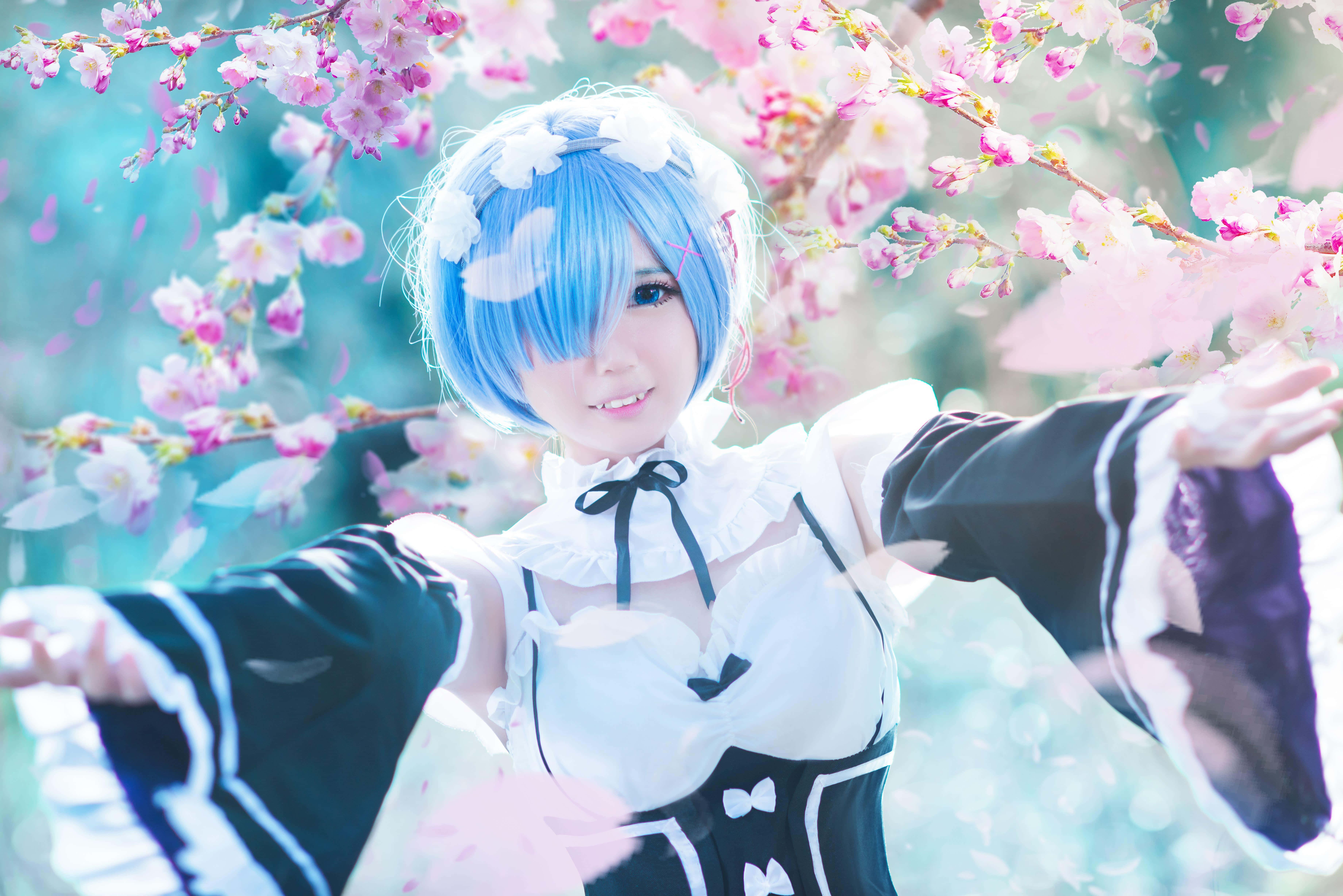 Cosplay Anime Kawaii 桌布 壁紙 — 一位藍色頭髮、身穿女僕裝的cosplayer站在盛開的粉紅色櫻花下，花瓣在她周圍飄落。