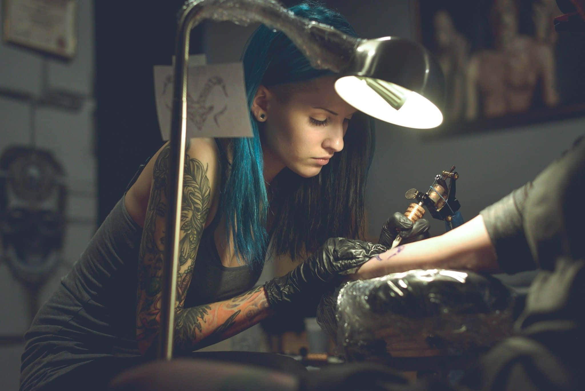 Tattoo 桌布 壁紙 — 一位藍髮、手臂有紋身的紋身師在昏暗的工作室中，專注地在明亮的燈光下為客戶的手臂紋身。