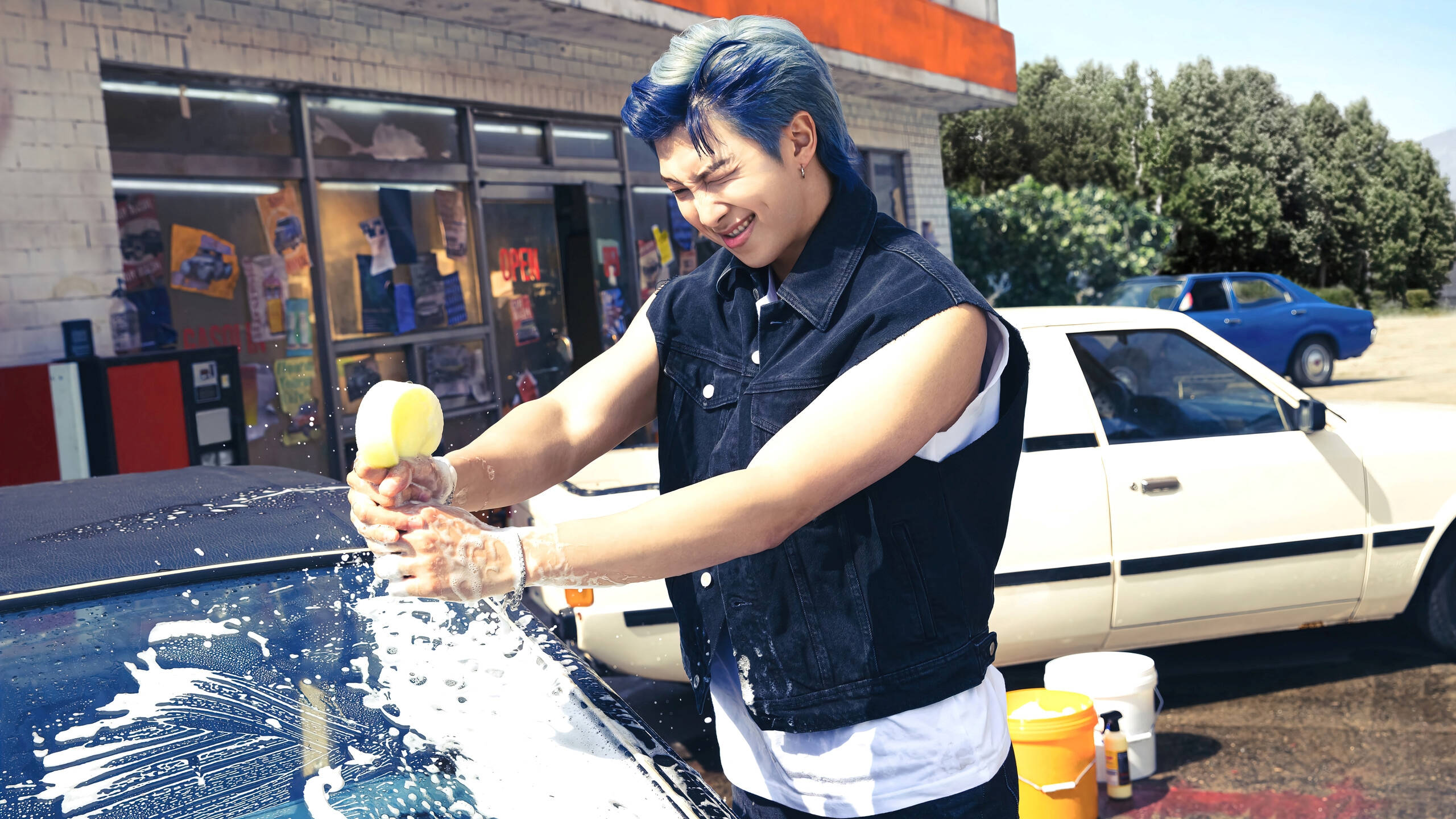 Wash 桌布 壁紙 — 一位藍髮年輕人在陽光明媚的洗車場愉快地清洗一輛深色汽車，肥皂泡飛揚。