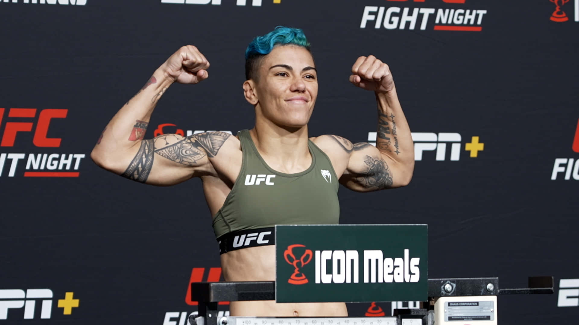 MMA 桌布 壁紙 — 一位藍髮的女性UFC拳手在明亮的燈光下於稱重活動中展示她帶有刺青的手臂。