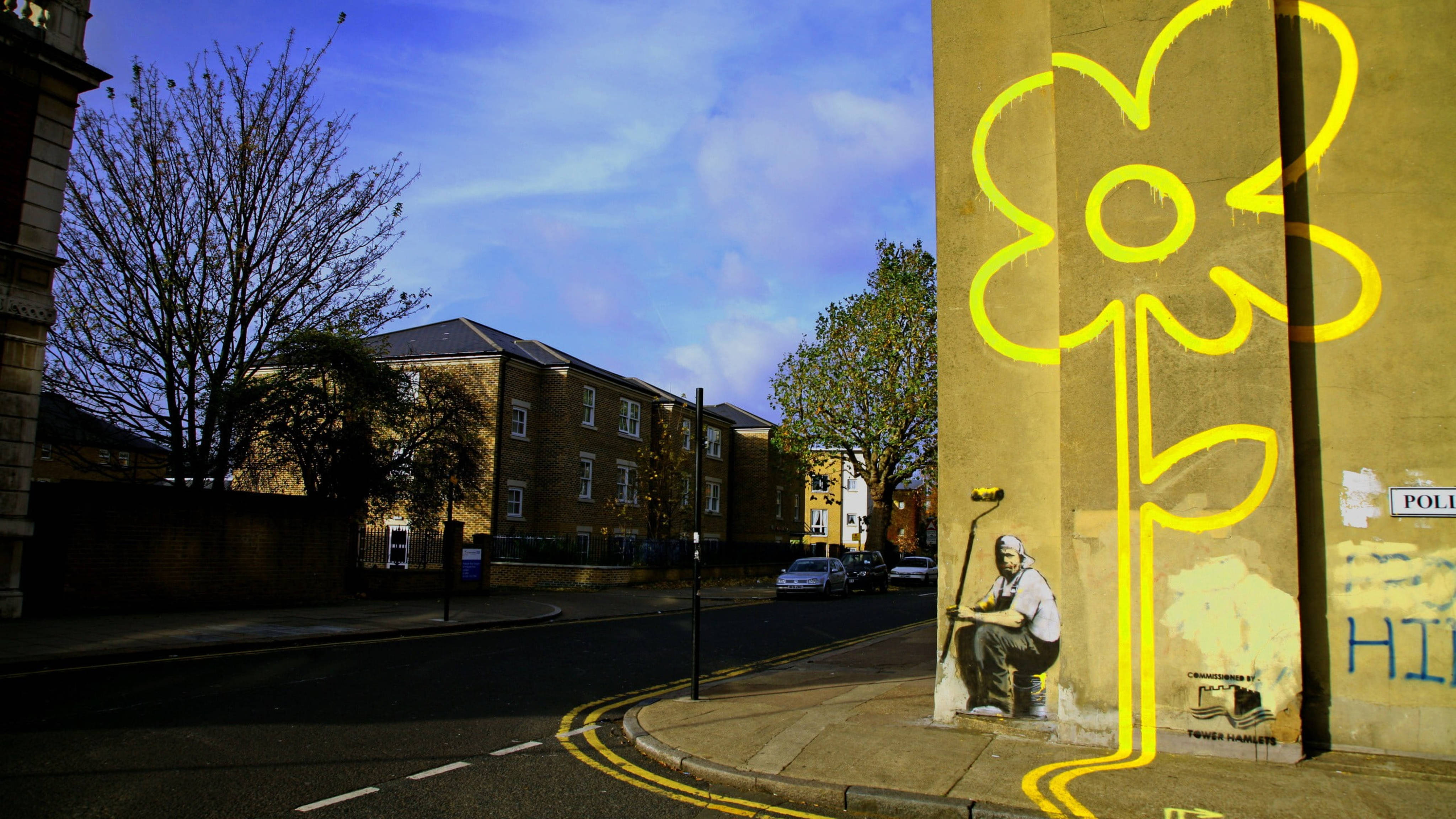Banksy 桌布 壁紙 — 一位街頭藝術家在藍天下，在一條寧靜的城市街道旁，有裸樹和停泊的汽車的磚牆上畫了一幅巨大的黃色花朵壁畫。