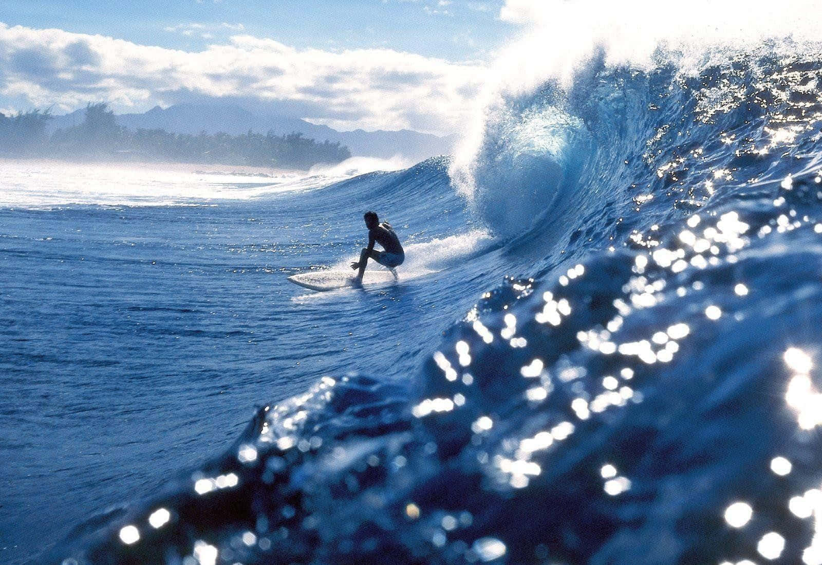 surfing 桌布 壁紙 — 一位衝浪者騎乘高聳的藍色海浪，陽光照射的水花四濺，遠山和陰天為海洋場景構框。