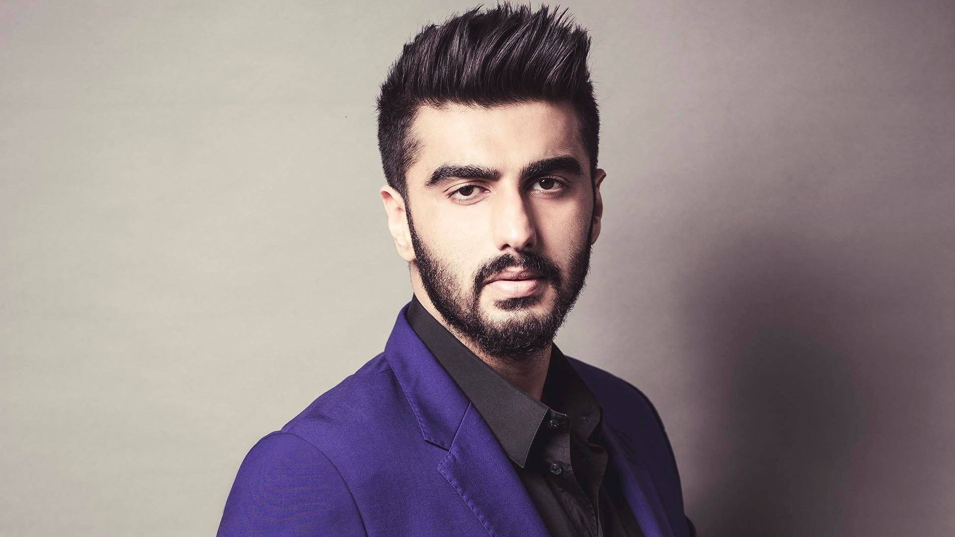 Arjun Kapoor 桌布 壁紙 — 一位衣著講究、留著時尚鬍鬚和尖刺頭髮的男士，在柔和的灰色工作室背景下穿著大膽的紫色西裝外套。