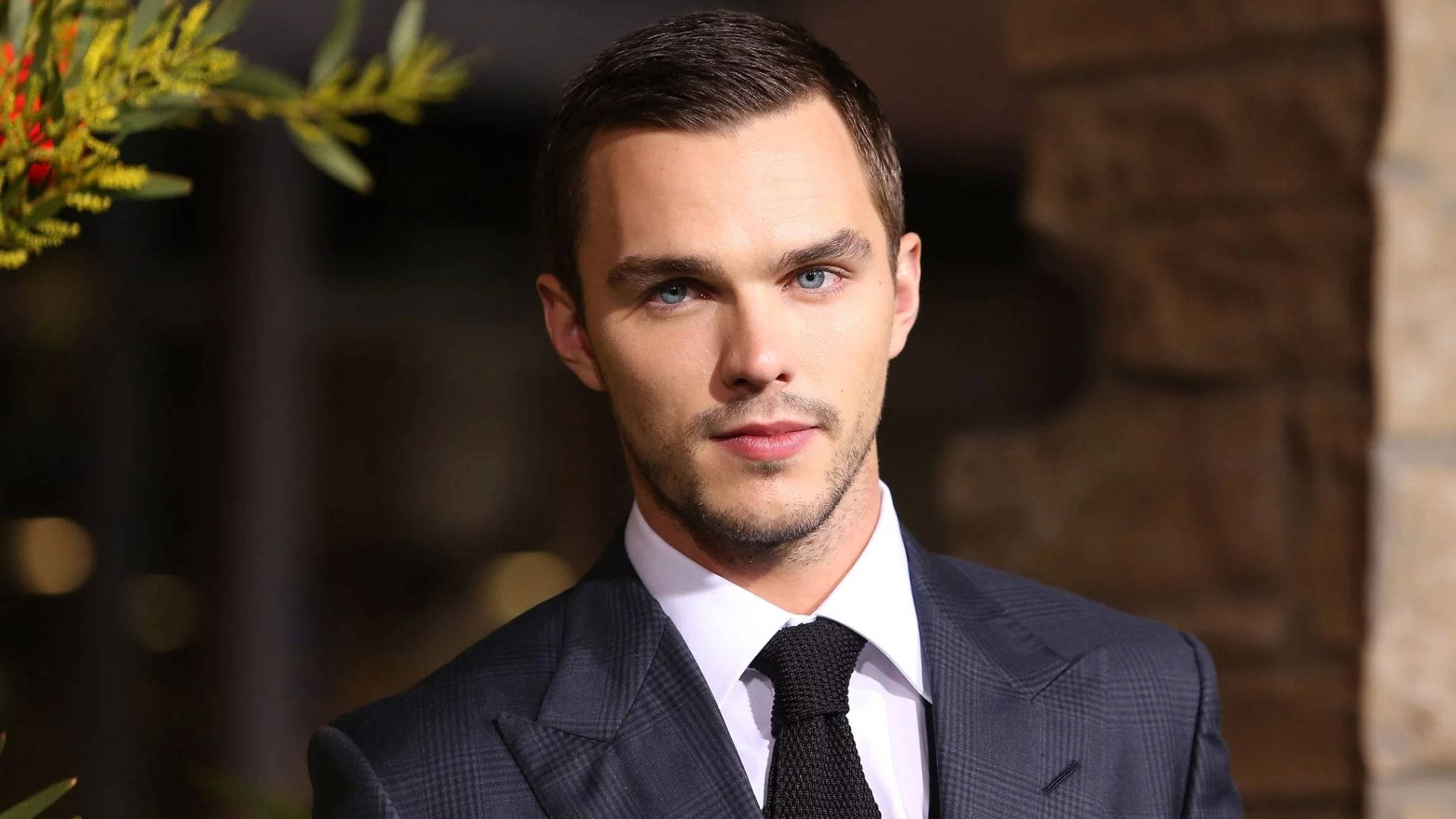 Nicholas Hoult 桌布 壁紙 — 一位衣著講究、身穿深色西裝打領帶的男士，在柔和模糊的石頭和樹葉背景下，用銳利的藍色眼睛直視鏡頭。