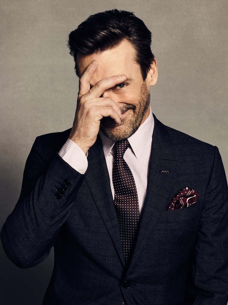 Jon Hamm 桌布 壁紙 — 一位衣著講究、身穿深色西裝的男子，在柔和的灰色背景下，頑皮地遮住一隻眼睛，露出得意的笑容。