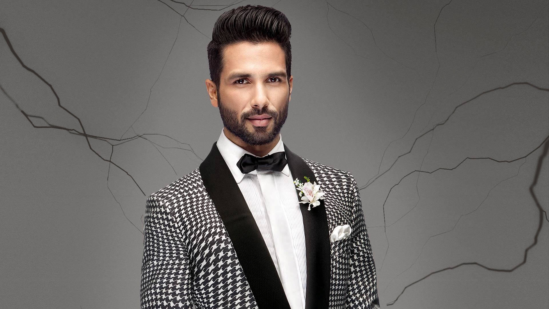 Shahid Kapoor 桌布 壁紙 — 一位衣著講究的男士身穿千鳥格禮服，佩戴白色襟花，站在一道裂縫的灰色牆前。