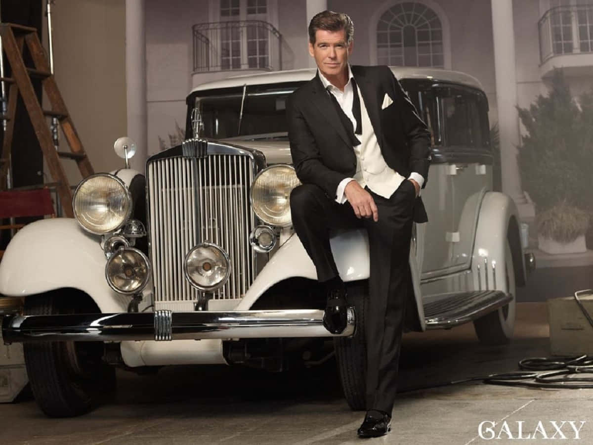 pierce brosnan 桌布 壁紙 — 一位衣著講究的男士身穿黑色西裝，在攝影棚內自信地靠在一輛復古白色豪華車旁。