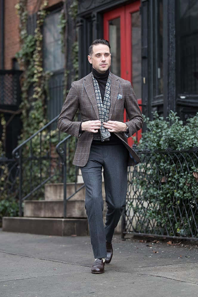 Man In Suit 桌布 壁紙 — 一位衣著講究的男子，身穿棕色格紋西裝外套和圖案圍巾，自信地走在城市人行道上，經過爬滿常春藤的建築物。