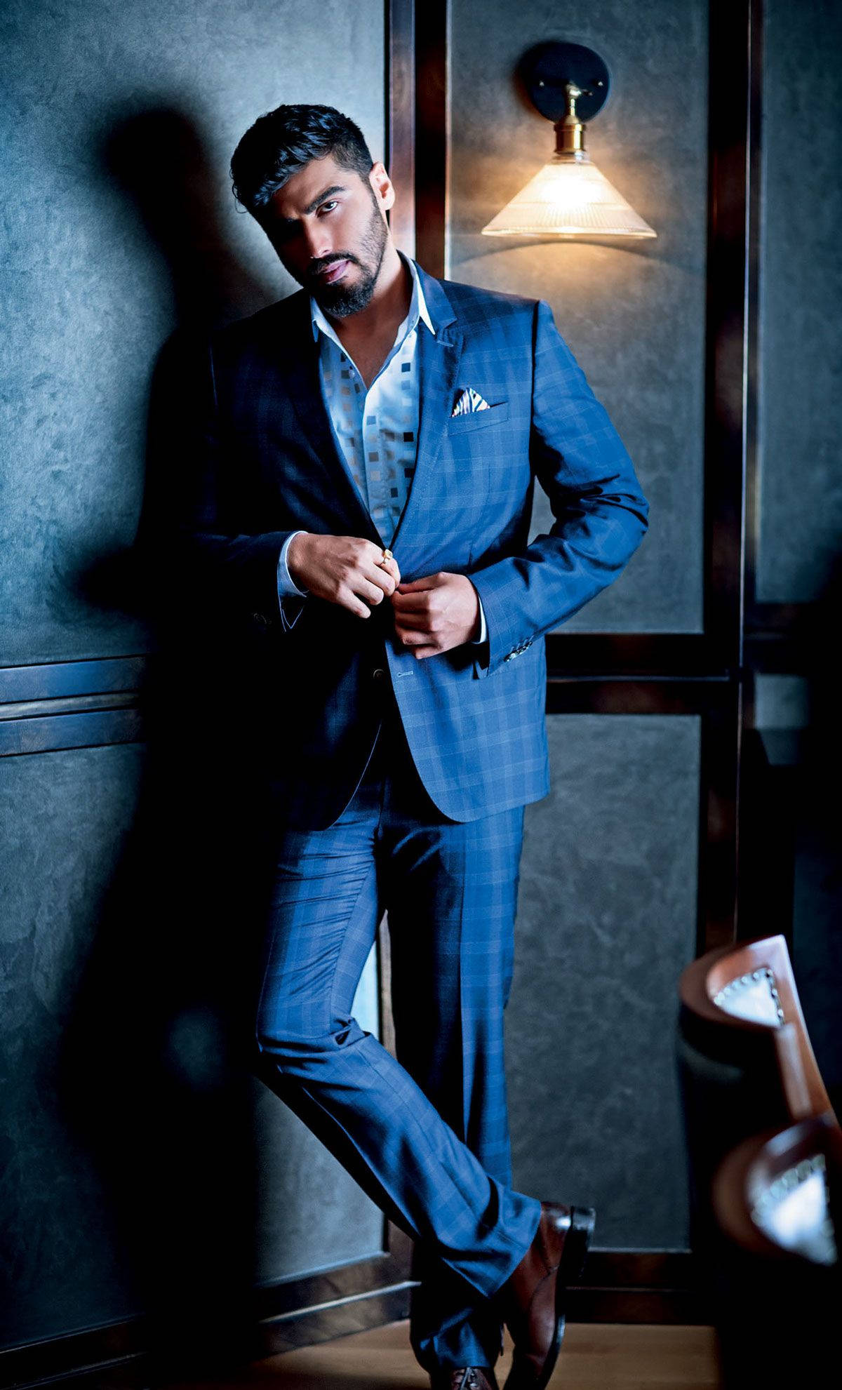 Arjun Kapoor 桌布 壁紙 — 一位衣著講究的男子身穿藍色格紋西裝，在溫暖的壁燈光線下，對著紋理灰色牆壁扣上外套鈕扣。