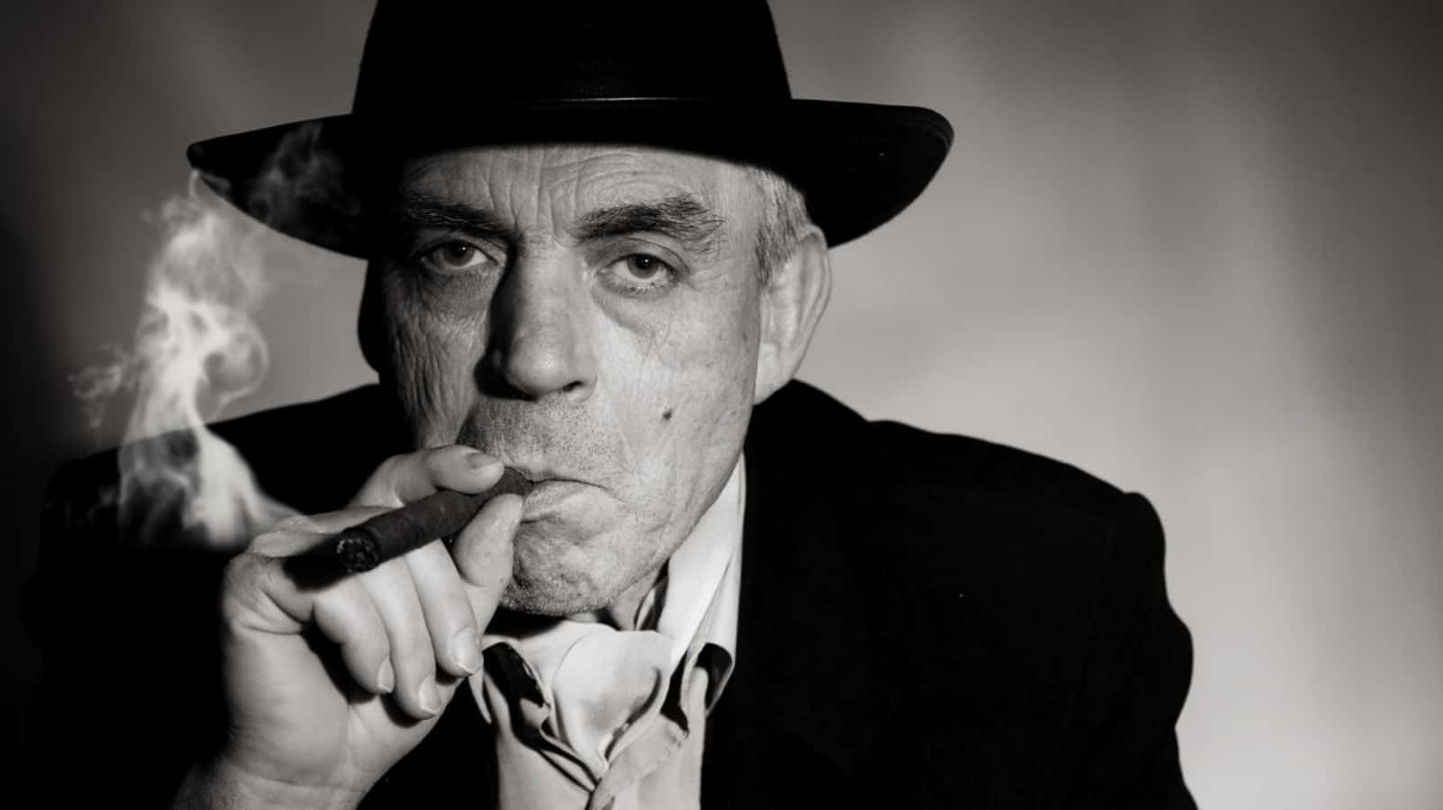 Mafia 桌布 壁紙 — 一位表情嚴肅、戴著紳士帽的年長男性抽著雪茄，煙霧從他的嘴唇繚繞升起，背景是柔和的灰色。