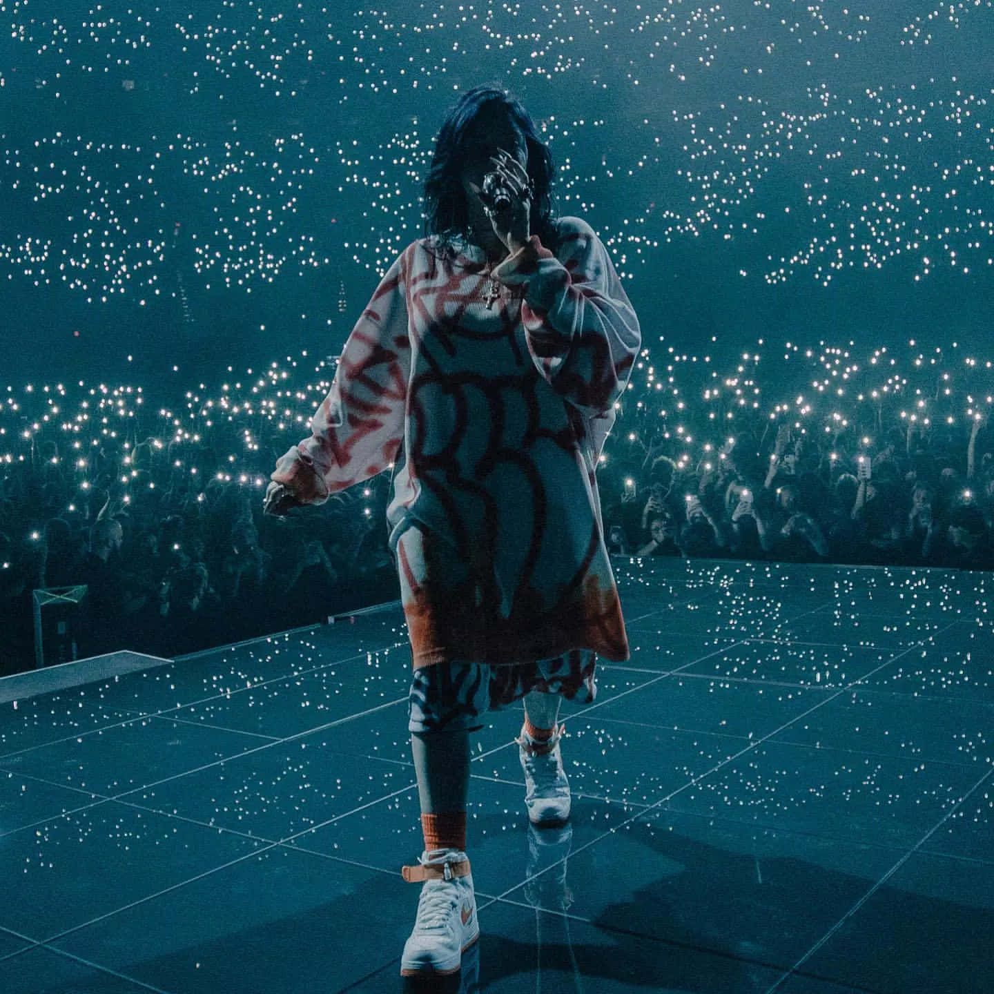 Billie Eilish 2021 桌布 壁紙 — 一位表演者在反射舞台上沐浴在藍光中對著麥克風唱歌，而人群舉起發光的手機像星星一樣。