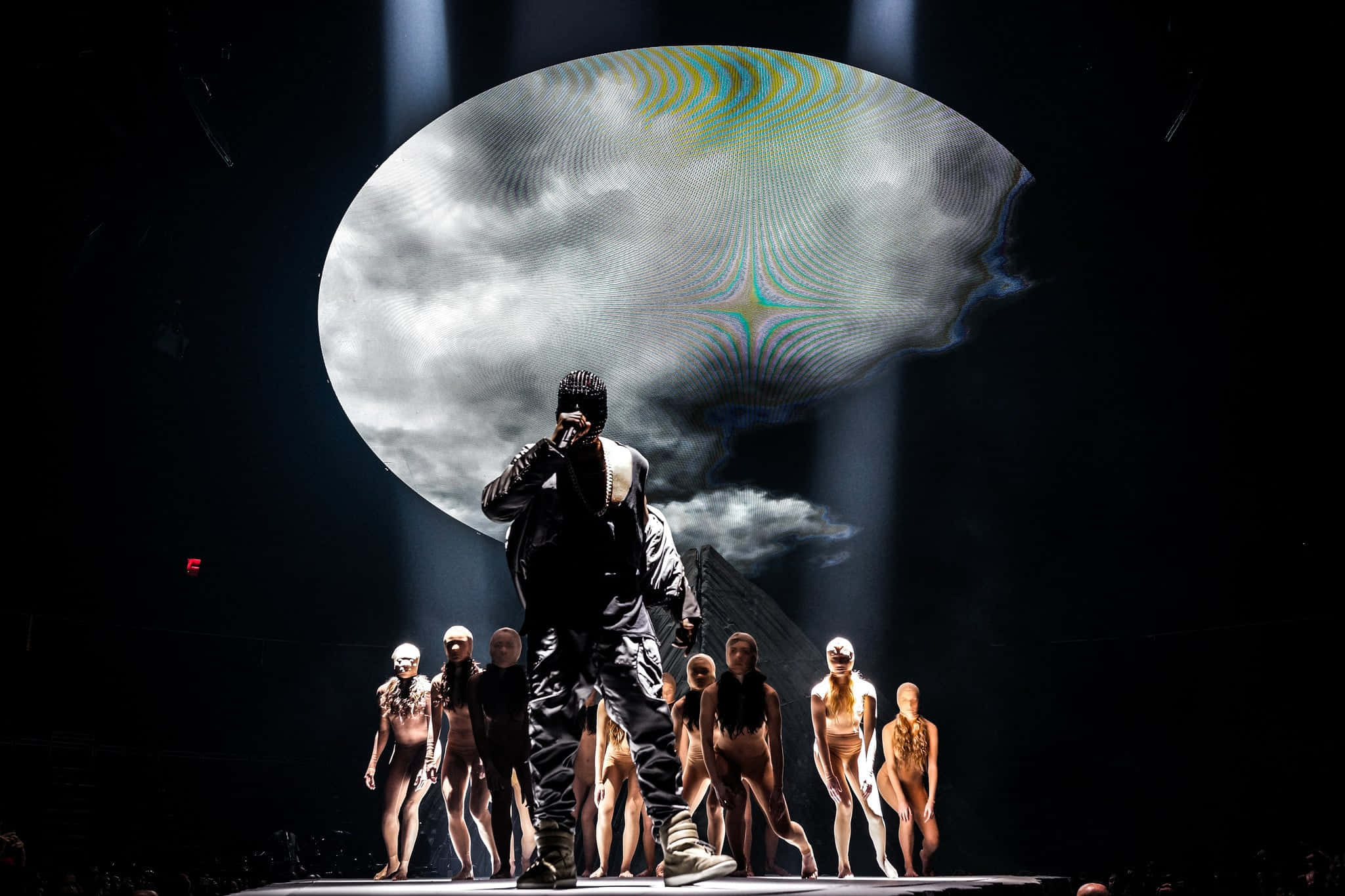 Kanye West Yeezus 桌布 壁紙 — 一位表演者在舞台上，處於戲劇性的聚光燈下，背後有超現實的雲狀投影，場地昏暗，有裸體舞者。
