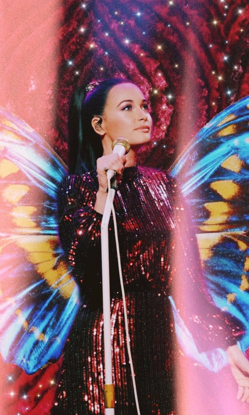 Kacey Musgraves 桌布 壁紙 — 一位表演者帶著蝴蝶翅膀，在星光紅燈下穿著閃閃發光的亮片服裝對著麥克風唱歌。