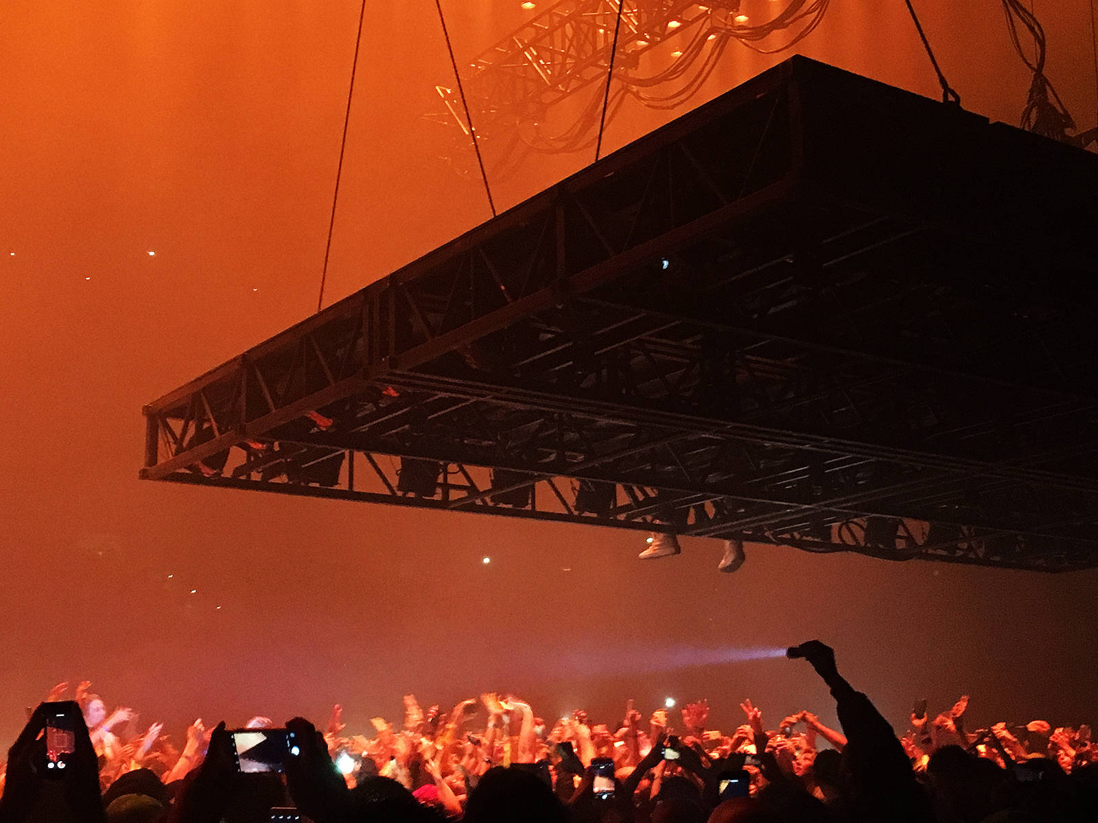 kanye west saint pablo 桌布 壁紙 — 一位表演者懸掛在懸掛式舞台裝置上，下方是沐浴在熾熱橙色演唱會燈光中的歡呼人群。