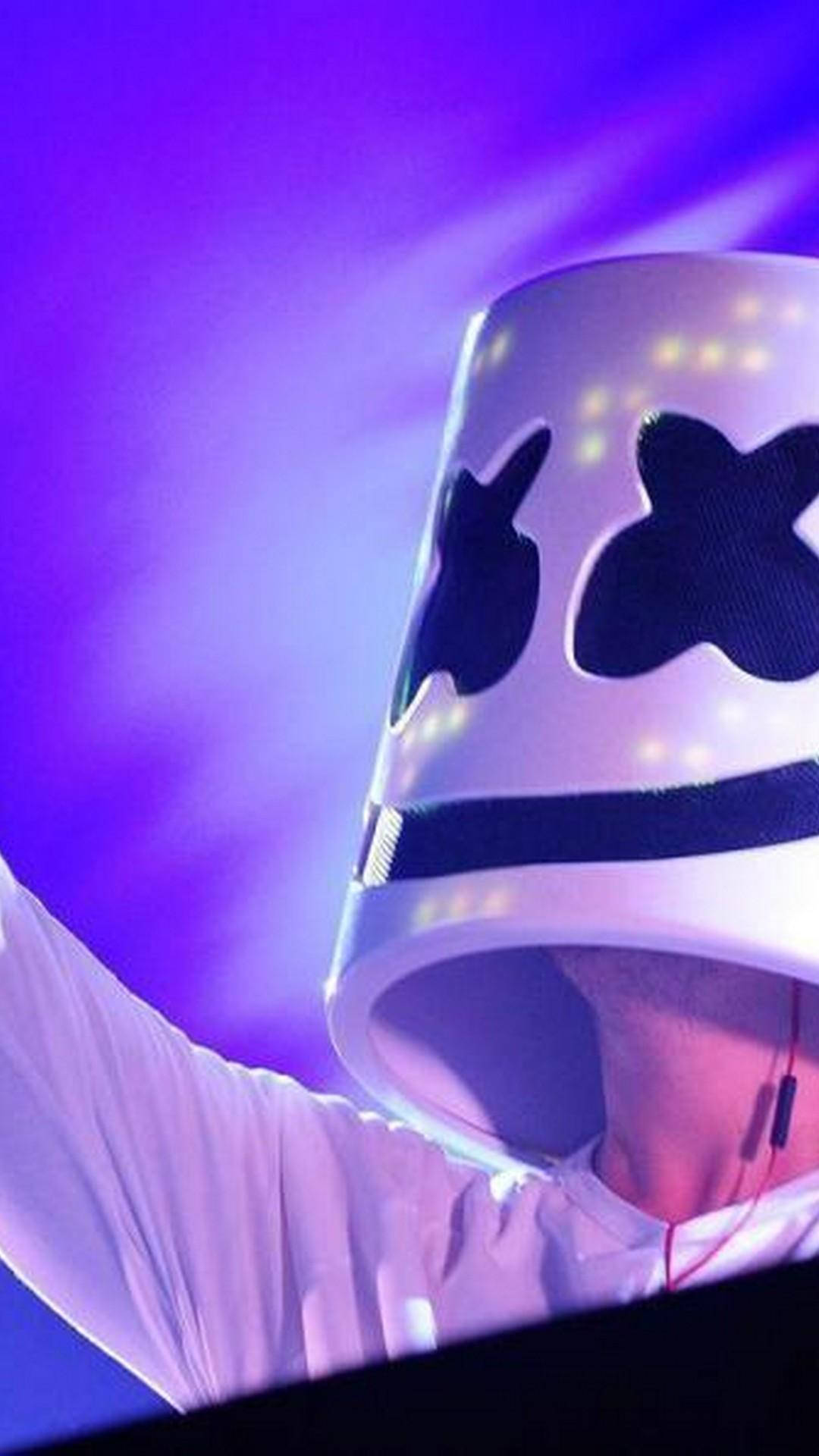marshmello iPhone 桌布 壁紙 — 一位表演者戴著發光的白色頭盔，帶有黑色X眼睛，在充滿活力的紫色舞台背景下舉起手臂。