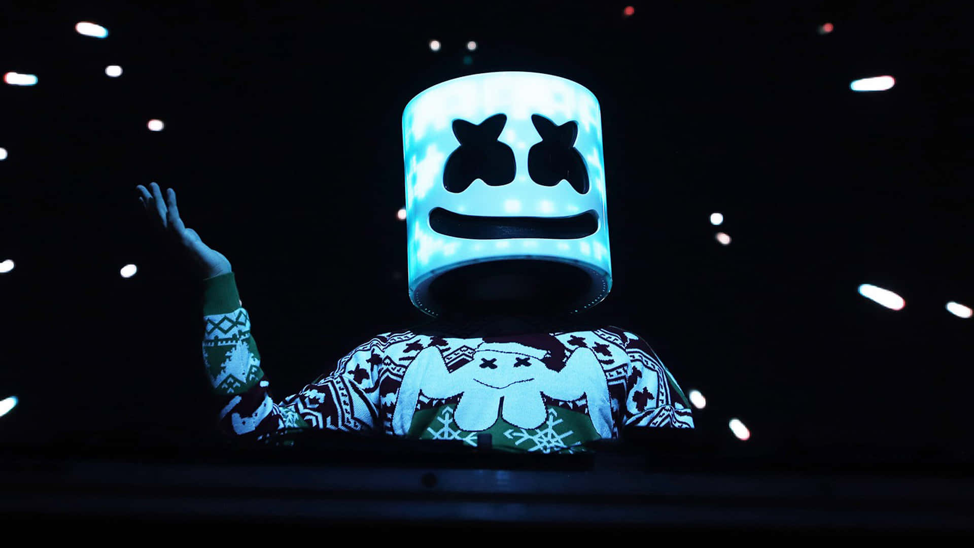 Marshmello 動態圖 桌布 壁紙 — 一位表演者戴著發光的藍色棉花糖頭盔和節日毛衣，在一個有漂浮燈光的黑暗演唱會舞台上揮手。