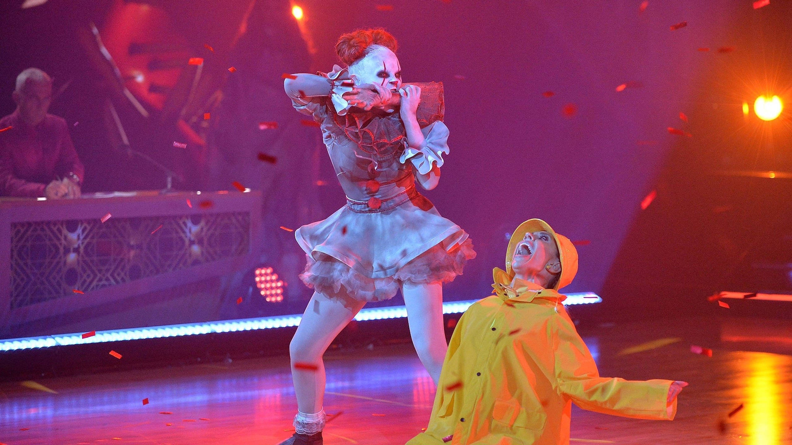JoJo Siwa 桌布 壁紙 — 一位表演者戴著白色骷髏面具和荷葉邊連衣裙，與另一位穿著黃色雨衣的表演者在充滿活力的舞台上對峙，五彩紙屑飄落。