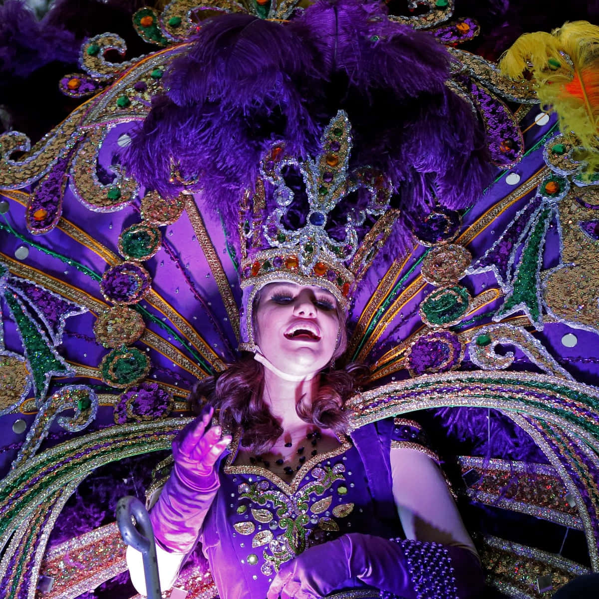 Mardi Gras 桌布 壁紙 — 一位表演者戴著耀眼的紫色羽毛頭飾和服裝，在充滿活力的舞台燈光下熱情歌唱。
