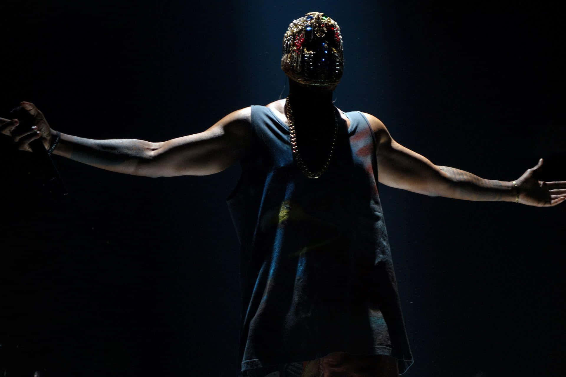 Kanye West Yeezus 桌布 壁紙 — 一位表演者戴著鑲有寶石的面具和背心，在黑色背景前，於戲劇性的舞台燈光下張開雙臂站立。