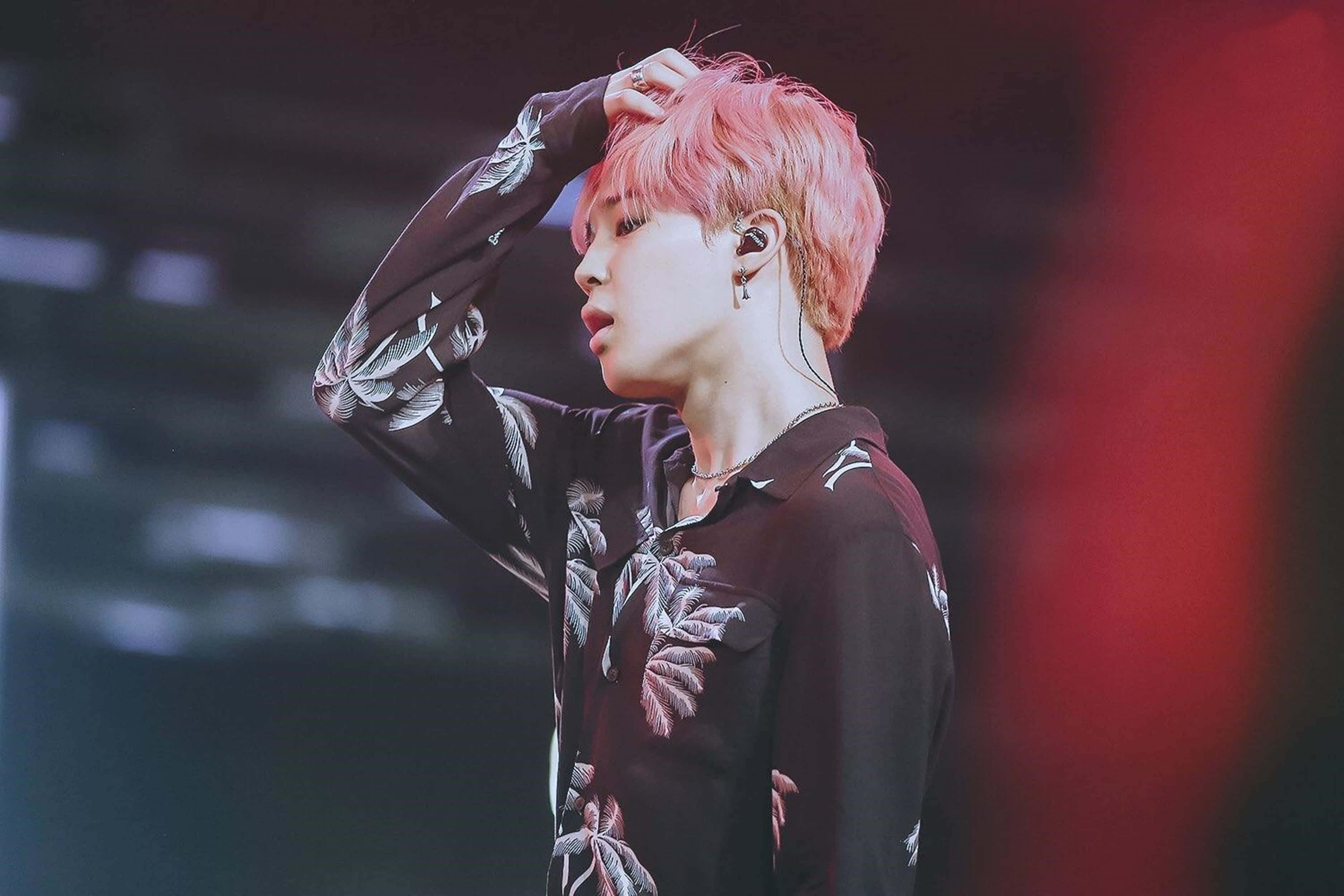 Jimin Aesthetic 桌布 壁紙 — 一位表演者擁有充滿活力的粉紅色頭髮和花卉襯衫，在舞台燈光下用手梳理頭髮。