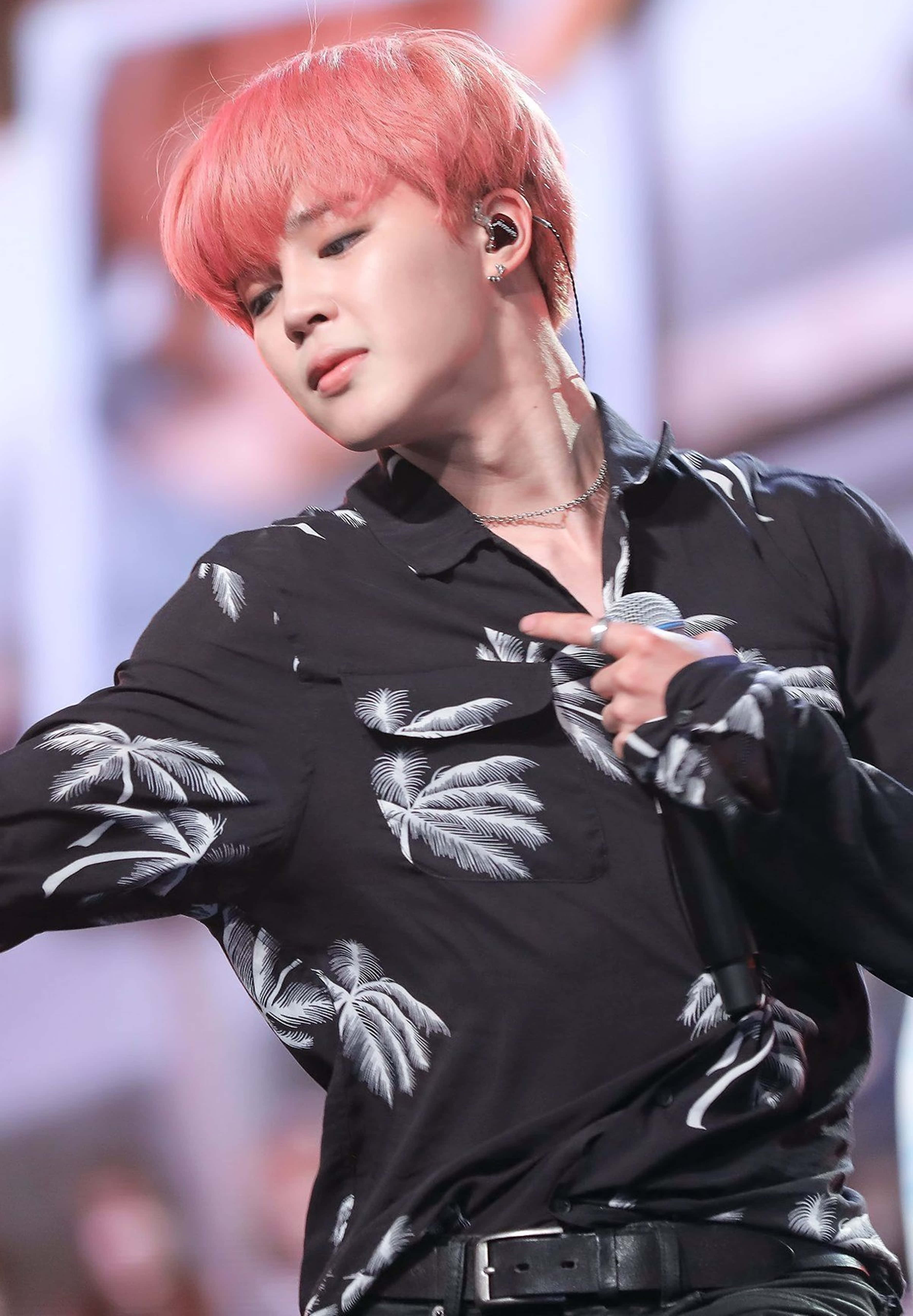 BTS 桌布 壁紙 — 一位表演者擁有充滿活力的粉紅色頭髮和黑色棕櫚樹襯衫，在明亮的燈光下在舞台上對著麥克風唱歌。