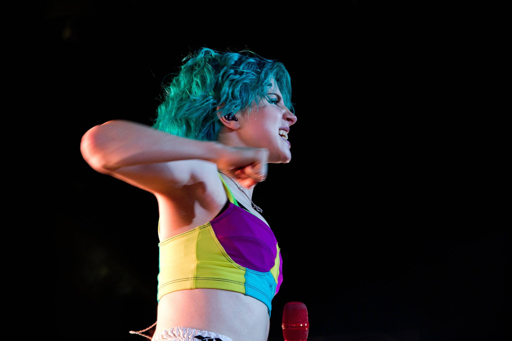 Hayley Williams 桌布 壁紙 — 一位表演者擁有充滿活力的藍綠色頭髮和彩虹短上衣，在黑色背景的舞台燈光下熱情歌唱。
