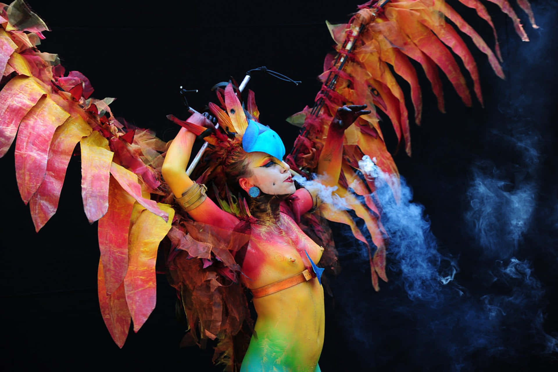 body art 桌布 壁紙 — 一位表演者擁有火紅橙色和黃色的羽毛翅膀，在黑色背景下呼出煙霧。