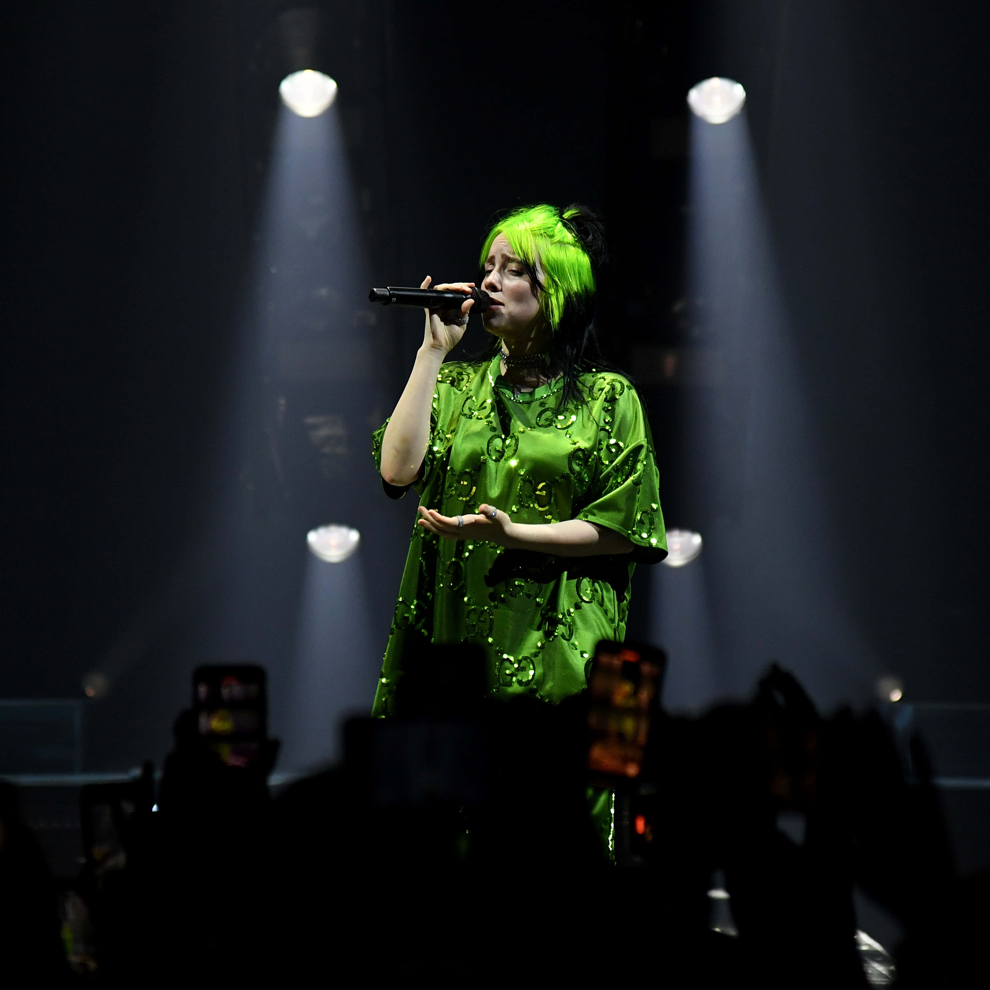 怪奇比莉 桌布 壁紙 — 一位表演者擁有螢光綠色頭髮和服裝，在明亮的舞台燈光下對著麥克風唱歌，下方可見觀眾的手機。