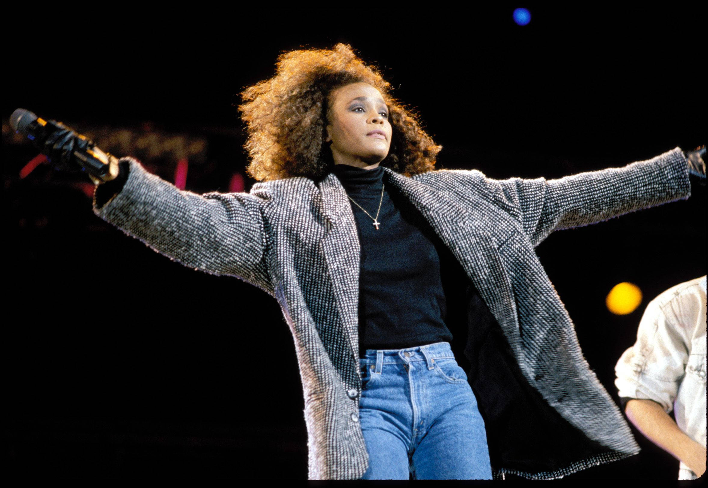 Whitney 桌布 壁紙 — 一位表演者擁有豐盈的捲髮，站在舞台上雙臂張開，手持麥克風，穿著質感外套和牛仔褲，在明亮的舞台燈光下。